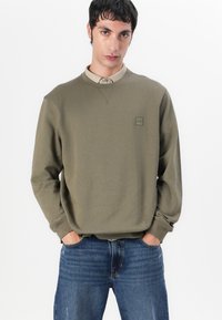 Olivengrønn hettegenser med ribbestrikkede mansjetter og rund hals, med et lite firkantet logopatch. Kombinert med blå denimjeans.