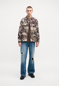 Multicolored patterned shirt featuring a branch and bird design, paired with light blue distressed jeans and black shoes.