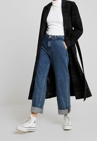 Persona che indossa jeans blu a maniche, sneaker bianche alte, dolcevita bianco e un lungo cappotto a quadri scuri con le mani nelle tasche.