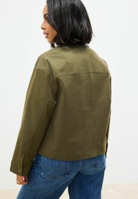 Dunkelgrüne Jacke mit standardmäßigem Kragen, langen Ärmeln und Knopfmanschetten, kombiniert mit blauen Jeans. Der Stoff wirkt glatt und strapazierfähig.