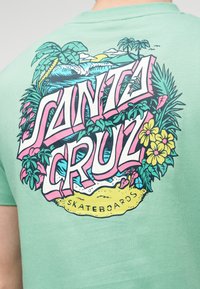 Πίσω μέρος πουκάμισου σε μέντα πράσινο με πολύχρωμο κείμενο «Santa Cruz Skateboards» περιτριγυρισμένο από τροπικά φυτά, φοινικόδεντρα, λουλούδια, κύματα και πουλιά.