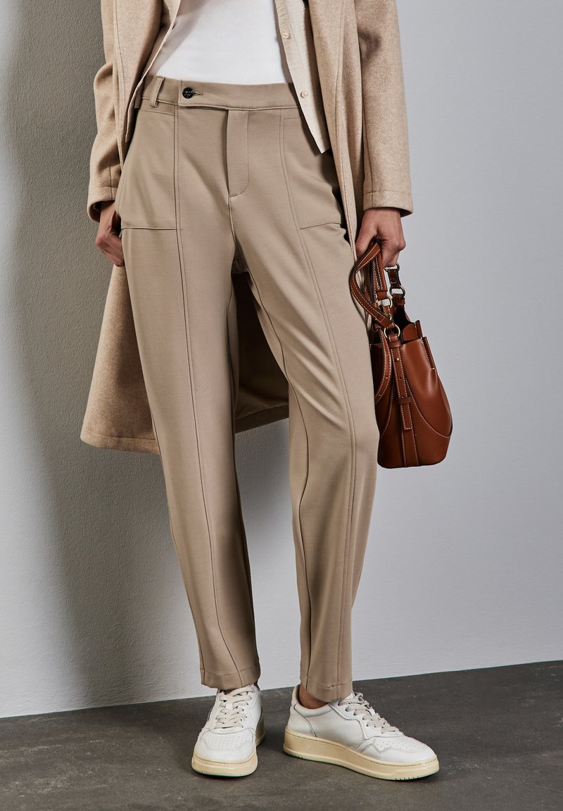 Beige tapered broek met voornaad, knoopsluiting en twee voorkoeken, gecombineerd met witte sneakers en een bruine handtas.