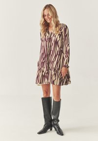 Robe à manches longues en violet et crème avec un motif en vagues, dotée d'un ourlet volanté et d'un nœud au col, associée à des bottes noires jusqu'au genou.