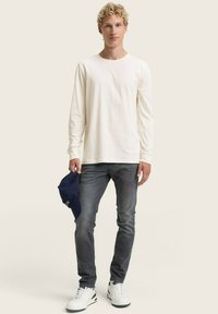 Junger Mann mit lockigem blonden Haar, der ein weißes Langarmshirt, graue Jeans und weiße Turnschuhe trägt und in seiner linken Hand eine marineblaue Mütze hält.