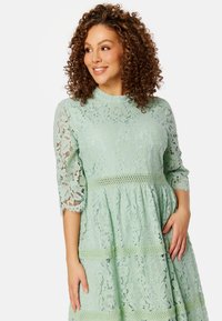 Abito in pizzo verde menta con maniche a tre quarti, scollatura alta, design a strati con dettagli in crochet e motivi floreali.