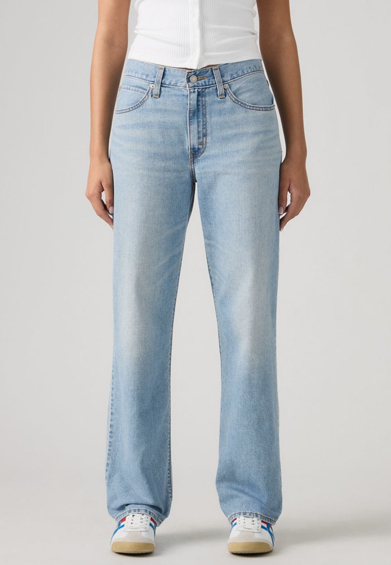 Γυναίκα που φοράει ανοιχτό μπλε τζιν με ίσια πόδια, λευκό ριμπ αμάνικο τοπ και λευκά αθλητικά παπούτσια με μπλε και κόκκινες ρίγες.