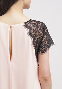 Haut en tissu rose avec des manches en dentelle noire aux bords festonnés. Détail au dos avec une ouverture en forme de goutte et une fermeture par bouton.