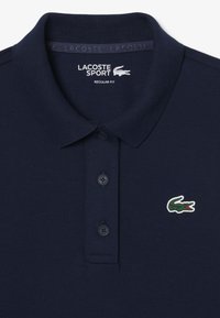 Marineblaues Poloshirt aus strukturiertem Stoff. Verfügt über einen klassischen Kragen, eine Dreiknopf-Leiste und ein grünes Krokodil-Logo auf der linken Seite.