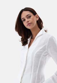 Camicetta di lino bianca con texture sottile, chiusura a bottoni e colletto. Presenta righe verticali e vestibilità su misura.