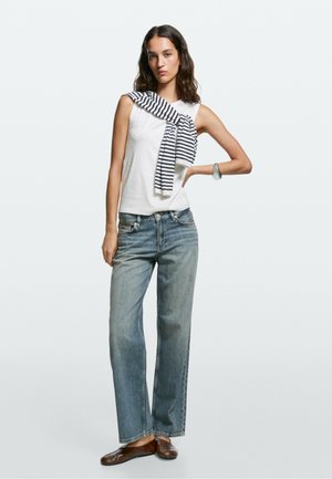 Junge Frau trägt ärmelloses weißes Top, gestreiften Pullover über der Schulter, blaue Jeans, braune flache Schuhe und Armband, steht mit einer Hand auf der Hüfte.
