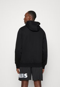 Sudadera con capucha negra de textura suave, que cuenta con un bolsillo tipo canguro y dobladillo acanalado. Lleva puesta con pantalones cortos de malla que exhiben letras reflectantes.