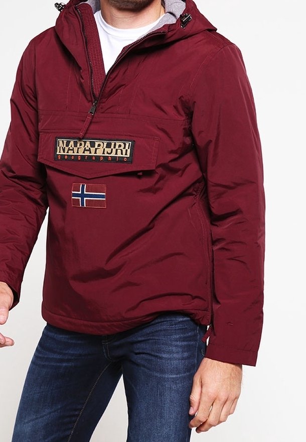 Man in een bordeauxrode Napapijri-jas met een voorste klepzak en Noorse vlagpatch, over een blauwe spijkerbroek, tegen een witte achtergrond.