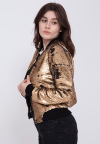 Goldfarbene Pailletten-Bomberjacke mit schwarzem geripptem Kragen und Bündchen, ausgestattet mit einer Reißverschlusstasche am Ärmel. Glatte Textur, taillierte Passform.