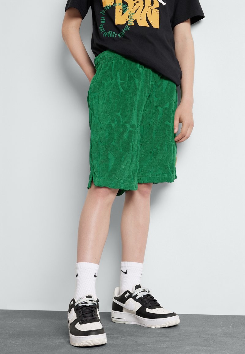 Personne portant un short vert texturé, des baskets noires et blanches, des chaussettes blanches Nike et un t-shirt graphique noir, debout contre un mur clair.