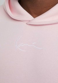 Närbild av en person som bär en ljusrosa huvtröja med en vit broderad signaturstil-logotyp på bröstet.