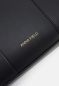 Černá kožená taška s vyraženým zlatým názvem značky "ANNA FIELD" na předním panelu, která se pyšní hladkým povrchem a detailním prošíváním.