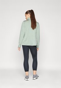 Maglietta verde chiaro a maniche lunghe con colletto alto, abbinata a leggings neri aderenti e scarpe da ginnastica grigie con dettagli testurizzati.