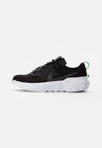 Black athletic sneaker with textured white sole, mesh upper, black strap over laces, and green pull tabs at heel and tongue.