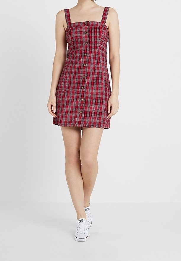 Vestito a quadri rosso e nero con scollatura quadrata, spalline regolabili e bottoni frontali. Indossato con sneakers bianche per un look casual.