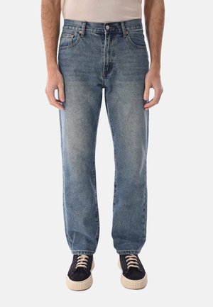 Uomo che indossa jeans blu slavati a gamba dritta e sneaker nere con suole bianche, in piedi con le braccia rilassate lungo i fianchi.