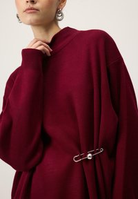 Burgundski oversized pulover z visokim ovratnikom, z dodatkom srebrne zaponke za varnost in rebrastimi manšetami za teksturo in podrobnosti.