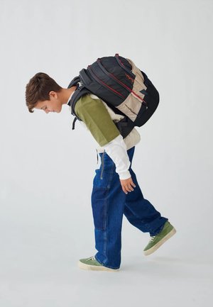 Schwarzer und beiger Rucksack mit roten Akzenten, verstellbaren Trägern und mehreren Fächern, getragen von einer Person in legerer Kleidung.