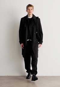 Schwarzer Trenchcoat mit doppelt geknöpften Knöpfen, über einem schwarzen Oberteil, kombiniert mit schwarzen Hosen und Turnschuhen. Glatte Textur, minimalistisches Design.