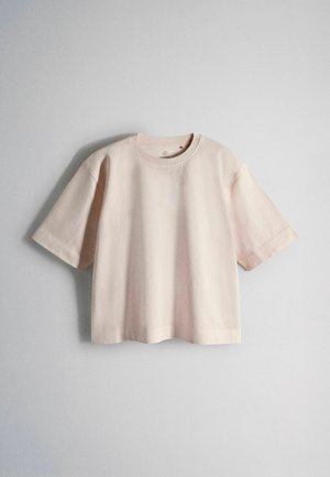 Tricou larg cu mâneci scurte și guler rotund, de culoare bej deschis, agățat pe un fundal simplu de perete gri deschis.