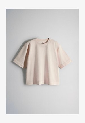 Tricou larg cu mâneci scurte și guler rotund, de culoare bej deschis, agățat pe un fundal simplu de perete gri deschis.