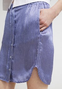 Ljuslila kjol med vertikala vita ränder, böjd fåll, knappar fram, elastisk midja och sidofickor. Slät textur.