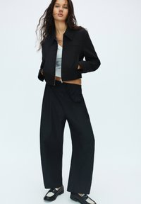 Jachetă neagră scurtă cu închidere cu fermoar și două buzunare frontale, purtată peste un top de culoare azul deschis și asortată cu pantaloni largi negri și pantofi negri.
