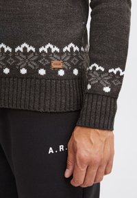 Dark gray knitted sweater with white snowflake patterns, ribbed cuffs, and a brown leather label. Worn with black pants featuring white lettering.