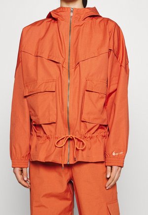Giacca arancione con cappuccio realizzata in tessuto leggero, con due ampie tasche frontali, vita regolabile con cordoncino e chiusura con zip.