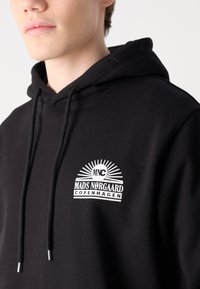 Sudadera negra con un logo frontal que presenta "MNC", "MADS NØRGAARD" y "COPENHAGUE". Diseñada con un cordón y textura suave.