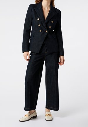 Femme portant un blazer sombre croisé avec des boutons dorés, un pantalon large sombre et des mocassins crème avec un détail chaîne dorée.