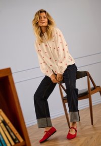 Vrouw met blond haar die een witte blouse met rode harten draagt, donkere opgerolde jeans en rode schoenen, zittend op een houten stoel binnen.