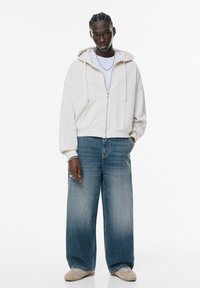 Felpa con zip di un leggero beige, dal fit rilassato, abbinata a jeans blu chiari a gamba larga e sneakers in tonalità neutra. Accessorio in catena argentata ben evidente.