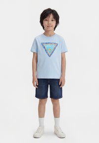 Αγόρι στέκεται μπροστά βλέποντας ευθεία, φορώντας ένα ανοιχτό γαλάζιο T-shirt Guess με λογότυπο, σκούρα τζιν σορτς, λευκές κάλτσες και λευκά αθλητικά παπούτσια σε απλό λευκό φόντο.