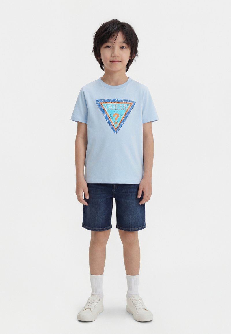 Αγόρι στέκεται μπροστά βλέποντας ευθεία, φορώντας ένα ανοιχτό γαλάζιο T-shirt Guess με λογότυπο, σκούρα τζιν σορτς, λευκές κάλτσες και λευκά αθλητικά παπούτσια σε απλό λευκό φόντο.