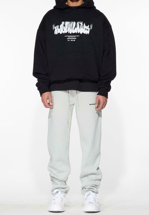 Person trägt einen schwarzen Hoodie mit weißem Grafiktext, hellgraue Jogginghosen mit kleinem schwarzem Logo am Oberschenkel und weiße Turnschuhe.