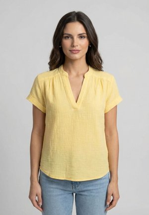 Femme aux cheveux foncés portant un chemisier jaune texturé à manches courtes et un jean bleu clair, debout devant un fond gris uni.