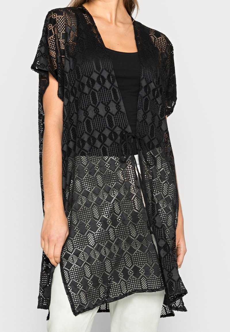 Femme portant un cardigan noir en crochet ouvert à l'avant avec des motifs géométriques par-dessus un haut noir et un pantalon blanc.