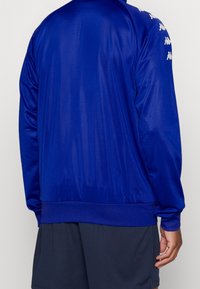 Blå sportjacka i slätt tyg med ribbade kanter vid nederkant och ärmslut. Har små logotypdetaljer längs ärmarna.