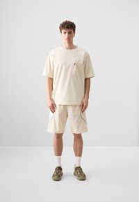 Beżowa koszulka z krótkim rękawem z kieszonką na piersi, zestawiona z beżowymi szortami z kontrastowymi szwami. Model ma na sobie oliwkowo-zielone sneakersy i białe skarpetki.
