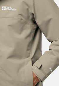 Giacca impermeabile beige con superficie testurizzata. Le caratteristiche includono un logo visibile, polsini regolabili e tasca laterale dal design elegante.