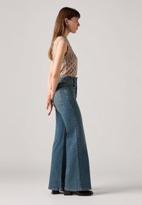 Calças de ganga de cintura alta e boca de sino em denim azul médio, apresentando um design de perna larga e detalhes de costura subtil, combinadas com um top de malha bege.