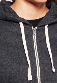 Šedá mikina na zip s texturovaným povrchem, vybavená smetanovými šňůrkami a kovovým zipem, zdůrazňuje měkkost látky.