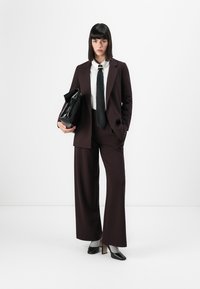 Completo su misura marrone scuro con pantaloni a gamba larga, camicia bianca, cravatta nera e borsa nera. Accompagnato da scarpe nere con tacco alto.