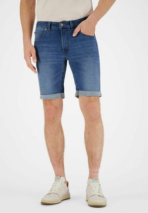 Man met opgerolde blauwe spijkershorts, witte sneakers en een wit T-shirt, staand met één hand in de zak op een witte achtergrond.