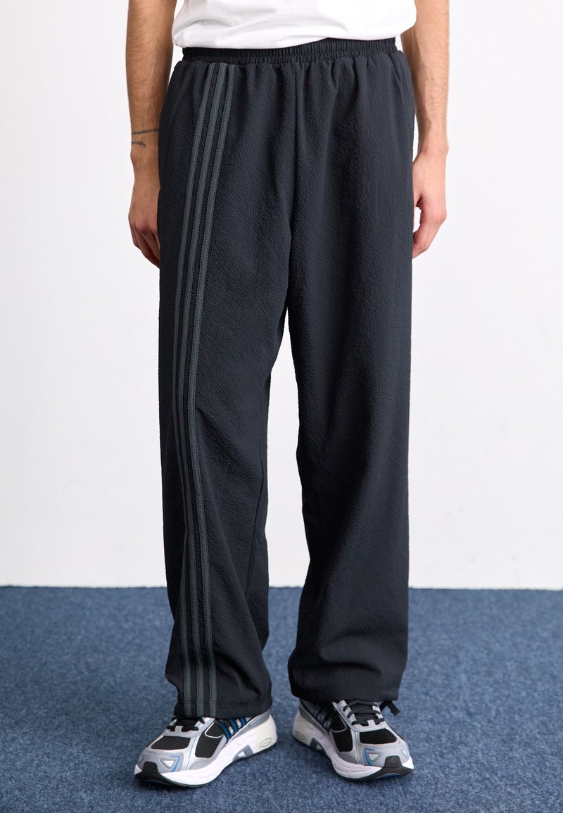 Svarta, texturerade träningsbyxor med elastisk midja och tre mörka ränder längs sidorna, kombinerade med grå och blå sneakers.